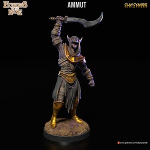 Könnte beinhalten: Eine bemalte Miniaturfigur von Ammut, einer Kreatur aus der ägyptischen Mythologie, die ein gebogenes Schwert hält. Die Figur ist in Grautönen, Braun und Gold bemalt und steht auf einem runden Sockel.
