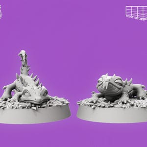 Könnte beinhalten: Zwei graue, 3D-gedruckte Fantasy-Kreaturen-Figuren. Eine steht aufrecht mit einem stacheligen Schwanz, die andere liegt. Beide stehen auf kreisförmigen Sockeln mit strukturierten Details, vor einem lila Hintergrund. Die Figuren sind wahrscheinlich für Tabletop-Spiele.