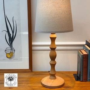 May include: A wooden table lamp with a light gray lampshade. The lamp has a turned wood base and a round wooden base. The lamp is next to a stack of books and a framed print.