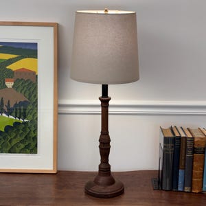 May include: A table lamp with a dark brown turned wood base and a cylindrical beige fabric lampshade. The lamp features a slender, vertically oriented stem with decorative molding details and a circular base, providing a neutral lighting fixture.