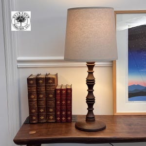 May include: A dark brown wooden table lamp with a beige lampshade. The lamp has a turned wood base and a decorative column. Several antique books are arranged on a wooden table, with a framed print in the background. The lamp is a decorative home decor item.