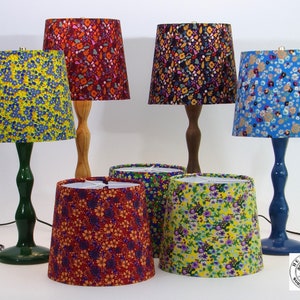 May include: Five table lamps with different floral patterned lampshades. The lampshades are in shades of blue, green, yellow, red, and brown. The lamps are on a white surface. The lamps are all different heights and have different colored bases.