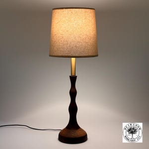 May include: A dark brown wooden table lamp with a beige lampshade. The lamp has a slender, curved stem and a round base with a textured design. The lampshade is a neutral color, and the lamp is lit.