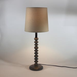 May include: A wooden table lamp with a beige lampshade. The lamp has a dark brown, beaded stem and a round base. A black power cord extends from the base. The lamp is on a white background.