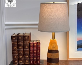 May include: A wooden table lamp with a beige lampshade. The lamp base features a layered design of light and dark brown wood. A collection of antique books is visible to the left of the lamp.