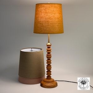 May include: A wooden table lamp with a tan lampshade. The lamp has a turned wood base and a brass stem. A second lampshade in tan and pink is next to the lamp. The lamp is a decorative home decor item.
