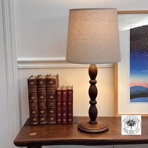 May include: A wooden table lamp with a beige lampshade. The lamp has a dark brown, turned wood base and stem. Several antique books with gold lettering and red covers are on the table. A framed print is visible in the background.