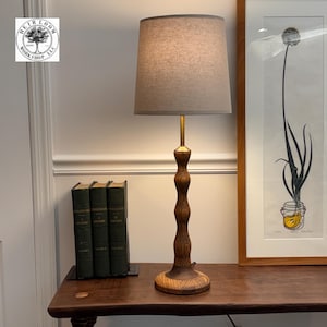 May include: A wooden table lamp with a brown, carved base and a beige lampshade. The lamp has a brass-colored stem. The lamp is on a wooden table with three green books and a framed picture in the background. The lamp is from Heirloom Workshop LLC.