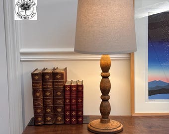 May include: A wooden table lamp with a light gray lampshade. The lamp has a turned wood base and a round wooden base. Several antique books are arranged on the table. A framed print hangs on the wall.