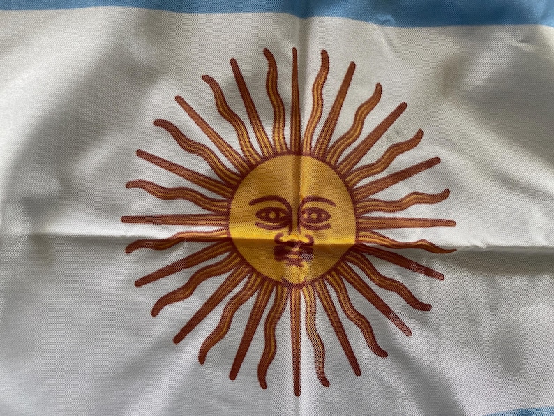 Puede incluir: Una bandera blanca con una franja azul en la parte superior y un gran sol naranja estilizado con una cara humana en el centro. El sol tiene una cara amarilla con ojos rojos y un bigote rojo.