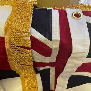 K&ouml;nnte beinhalten: Nahaufnahme einer rot-wei&szlig;-blauen Flagge mit goldenen Fransen. Die Flagge hat eine Messing&ouml;se in der oberen rechten Ecke.