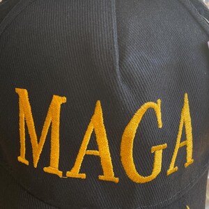 May include: Black baseball cap with the embroidered word "MAGA" in gold thread. The cap also features a gold embroidered signature on the side. The cap has a curved brim and an adjustable strap.
