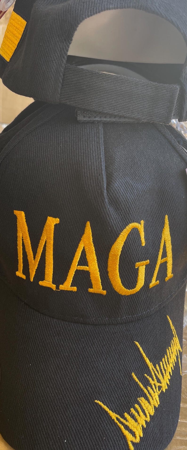 May include: Black baseball cap with the embroidered word "MAGA" in gold thread. The cap also features a gold embroidered signature on the side. The cap has a curved brim and an adjustable strap.