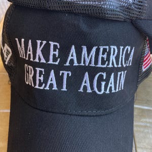 May include: Black baseball cap with the phrase "MAKE AMERICA GREAT AGAIN" embroidered in white. The cap has a mesh back and the numbers "45-47" embroidered in white on the mesh.