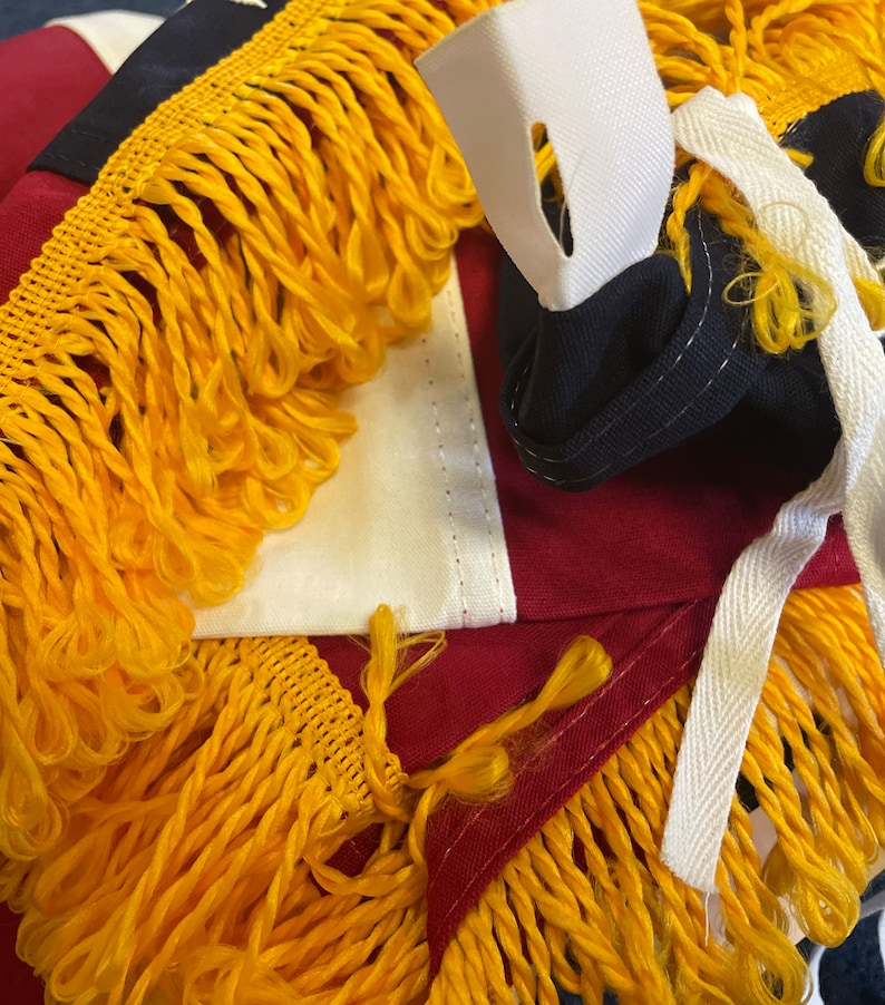 K&ouml;nnte beinhalten: Nahaufnahme einer rot-wei&szlig;-blauen amerikanischen Flagge mit goldenen Fransen. Die Flagge ist gefaltet und hat einen wei&szlig;en Riemen mit einem Loch.