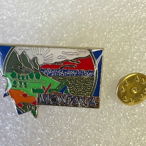 May include: A colorful enamel pin depicting the state of Montana. The pin features a stylized outline of the state with mountains, a sun, and the word "MONTANA" in white lettering. The pin is attached to a gold-colored metal backing.