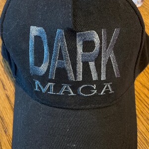 May include: Black baseball cap featuring the embroidered words "DARK MAGA" in light blue. The cap has a curved peak and a structured crown, suitable for casual wear.