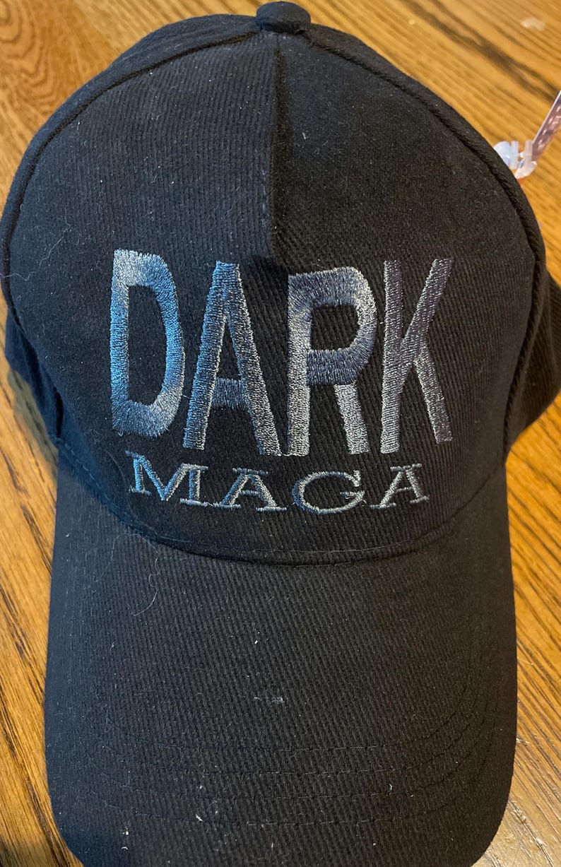 May include: Black baseball cap featuring the embroidered words "DARK MAGA" in light blue. The cap has a curved peak and a structured crown, suitable for casual wear.