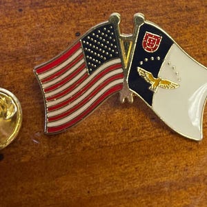 May include: A gold-colored pin with two flags. The flag on the left is the American flag with red and white stripes and blue stars. The flag on the right is the flag of Portugal with a white background, a blue shield with a red cross, and a gold eagle.