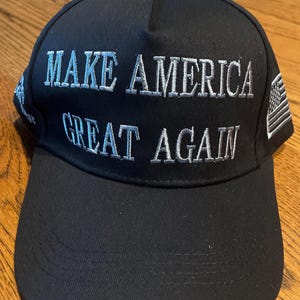 May include: Black baseball cap with the embroidered text "MAKE AMERICA GREAT AGAIN" in white. The cap features a curved brim and an American flag patch on the side. A classic, adjustable hat.