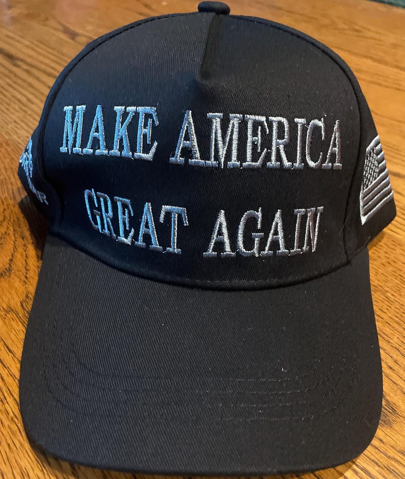 May include: Black baseball cap with the embroidered text "MAKE AMERICA GREAT AGAIN" in white. The cap features a curved brim and an American flag patch on the side. A classic, adjustable hat.