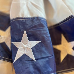 Puede incluir: Primer plano de una secci&oacute;n de una bandera con una estrella blanca bordada sobre un fondo azul marino. La bandera tiene rayas blancas y azul marino. La estrella es una estrella de cinco puntas.