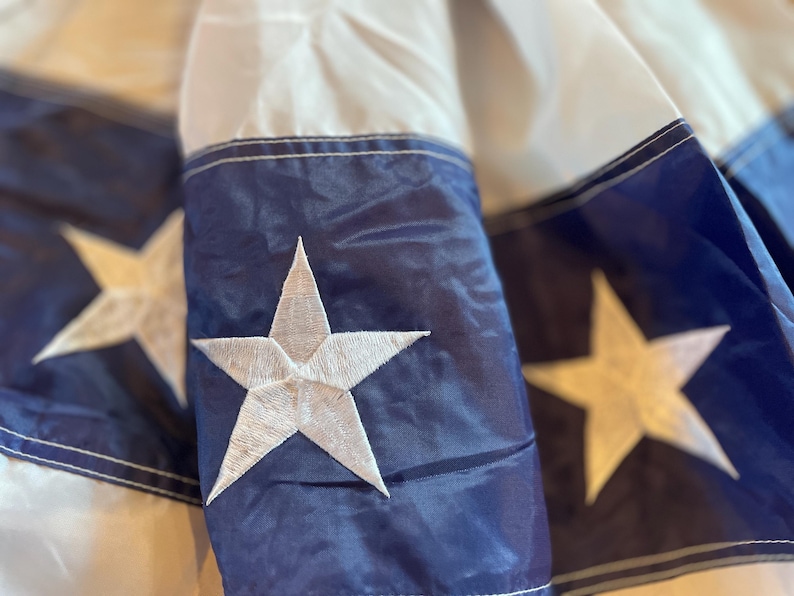 Puede incluir: Primer plano de una secci&oacute;n de una bandera con una estrella blanca bordada sobre un fondo azul marino. La bandera tiene rayas blancas y azul marino. La estrella es una estrella de cinco puntas.