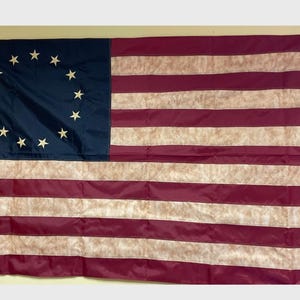 May include: A vintage-style American flag with 13 red and white stripes and a blue canton featuring 13 white stars in a circle. The flag has a faded, distressed look, giving it a vintage feel.