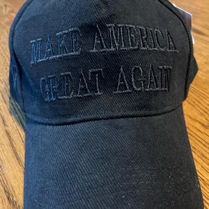 May include: Black baseball cap featuring the embroidered phrase "MAKE AMERICA GREAT AGAIN". The cap has a curved peak and a button on the crown. The hat is made of a textured material.