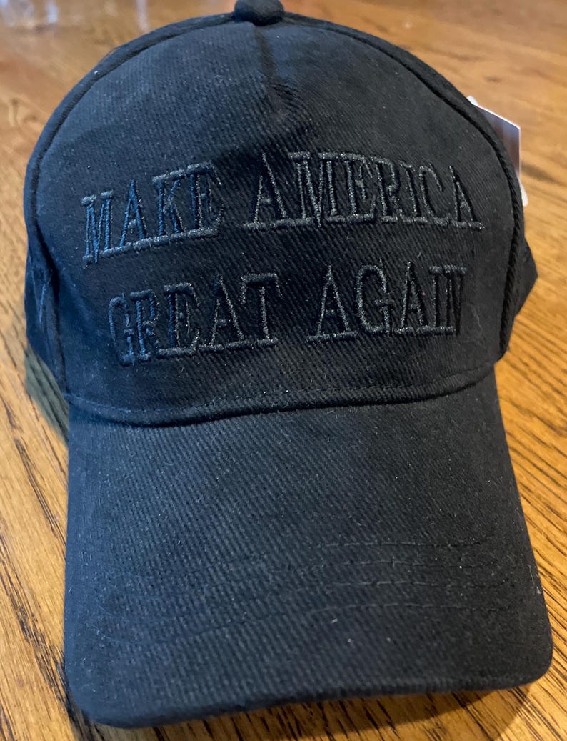 May include: Black baseball cap featuring the embroidered phrase "MAKE AMERICA GREAT AGAIN". The cap has a curved peak and a button on the crown. The hat is made of a textured material.