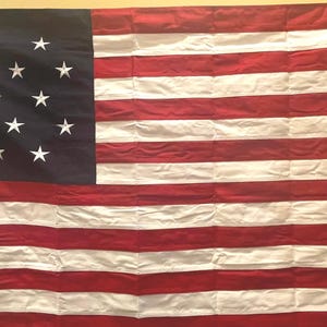 May include: A vintage American flag with 13 red and white stripes and a blue field with 13 white stars. The flag is made of fabric and has a slightly wrinkled appearance.