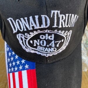 May include: Black baseball cap with the text "DONALD TRUMP" in white script above a white and black emblem that reads "old NO.47 BRAND". The left side of the cap features the American flag.