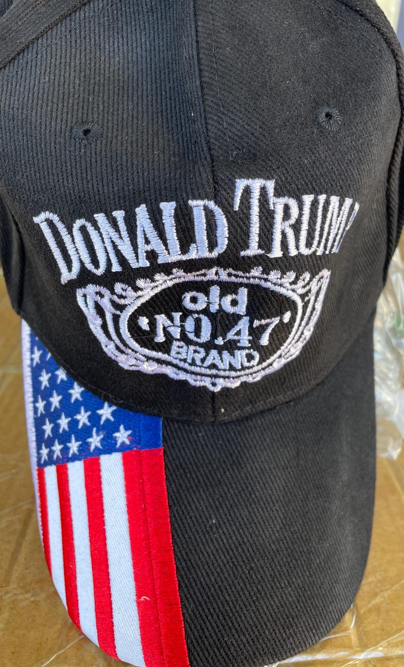 May include: Black baseball cap with the text "DONALD TRUMP" in white script above a white and black emblem that reads "old NO.47 BRAND". The left side of the cap features the American flag.
