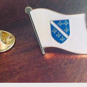 K&ouml;nnte beinhalten: Ein Ansteck-Pin mit der Flagge von Bosnien und Herzegowina. Der Pin hat einen wei&szlig;en Hintergrund mit einem blauen Schild mit sechs gelben Lilien. Der Pin ist aus Metall und hat einen goldfarbenen Verschluss.