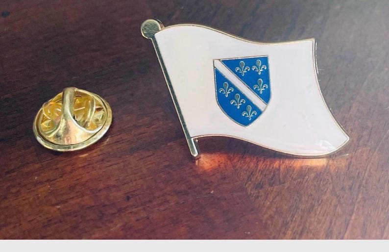 K&ouml;nnte beinhalten: Ein Ansteck-Pin mit der Flagge von Bosnien und Herzegowina. Der Pin hat einen wei&szlig;en Hintergrund mit einem blauen Schild mit sechs gelben Lilien. Der Pin ist aus Metall und hat einen goldfarbenen Verschluss.