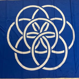 Puede incluir: Bandera azul con un diseño circular blanco entrelazado. Una franja blanca recorre el borde izquierdo. El diseño consta de seis círculos entrelazados, formando un patrón central en forma de flor. La bandera está hecha de un material ligero.