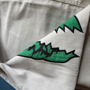 May include: A white canvas fabric with a green and black embroidered design of a jagged tooth shape.