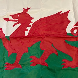 May include: A flag of Wales, featuring a red dragon centered on a white and green background. The dragon is detailed with wings, claws, and a long tail. The flag is made of a smooth, possibly nylon, material.