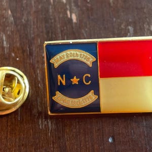 Puede incluir: Un pin rectangular de esmalte con el dise&ntilde;o de la bandera del estado de Carolina del Norte. El pin tiene un borde dorado y un respaldo dorado. El dise&ntilde;o de la bandera incluye una secci&oacute;n roja y dorada, y una secci&oacute;n azul con las letras "NC" y las fechas "May 20th 1775" y "April 12th 1776".