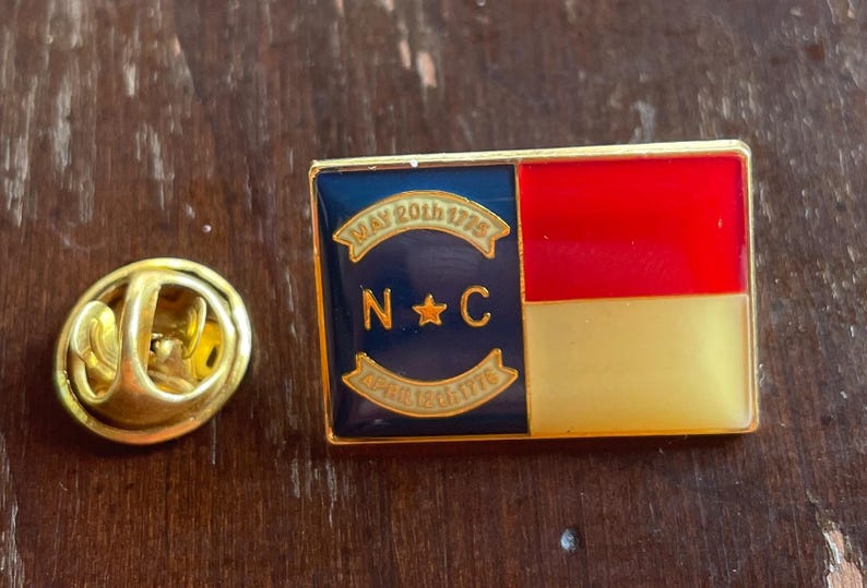 Puede incluir: Un pin rectangular de esmalte con el dise&ntilde;o de la bandera del estado de Carolina del Norte. El pin tiene un borde dorado y un respaldo dorado. El dise&ntilde;o de la bandera incluye una secci&oacute;n roja y dorada, y una secci&oacute;n azul con las letras "NC" y las fechas "May 20th 1775" y "April 12th 1776".