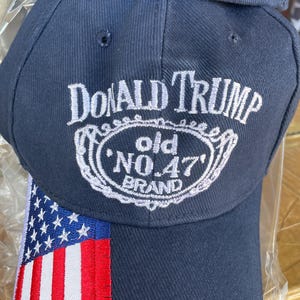 May include: Navy blue baseball cap with the text "DONALD TRUMP" above a white oval design with "old NO. 47 BRAND". The cap features an American flag design on the side.