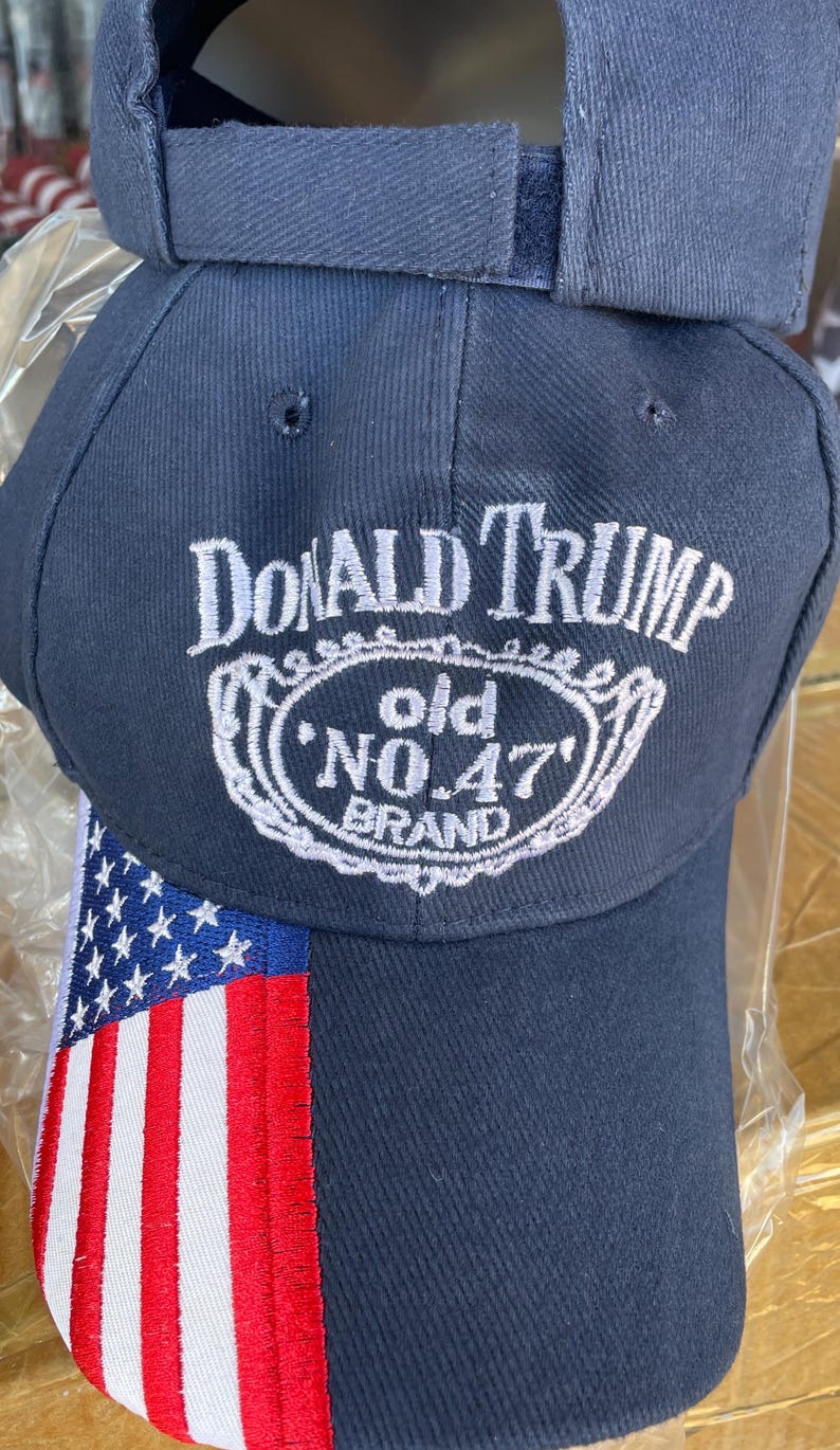 May include: Navy blue baseball cap with the text "DONALD TRUMP" above a white oval design with "old NO. 47 BRAND". The cap features an American flag design on the side.