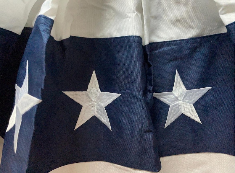 Puede incluir: Primer plano de una tela con tem&aacute;tica patri&oacute;tica. El dise&ntilde;o presenta estrellas blancas bordadas sobre un fondo azul marino, con rayas blancas y azul marino. La tela es probablemente parte de una bandera o un art&iacute;culo decorativo.