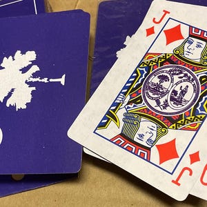 May include: A deck of playing cards featuring the Jack of Diamonds and the Queen of Diamonds. The cards have a white background with red and black details. The backs of the cards are blue with a white palmetto tree and crescent moon design.