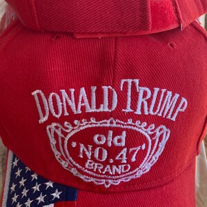 May include: Red baseball cap with the text "DONALD TRUMP" in white script above a white emblem with "old NO.47 BRAND". The cap has an American flag design on the side and an adjustable strap.
