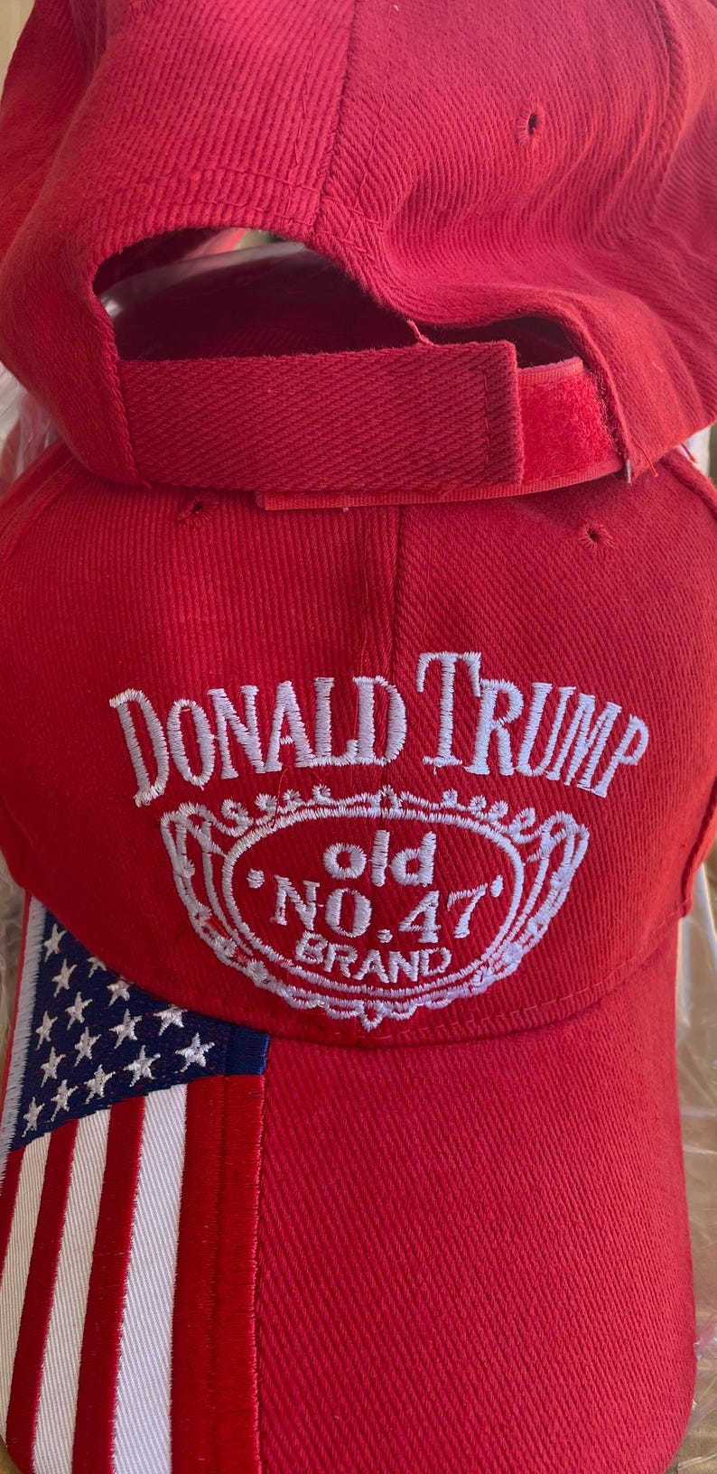 May include: Red baseball cap with the text "DONALD TRUMP" in white script above a white emblem with "old NO.47 BRAND". The cap has an American flag design on the side and an adjustable strap.