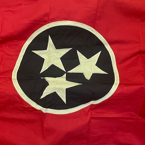 May include: A red flag with a central emblem featuring three white stars within a black circle, outlined in white. The flag is made of a fabric material and is likely a state flag.