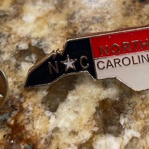 Puede incluir: Un pasador dorado y un pasador con la forma del estado de Carolina del Norte. El pasador presenta el contorno del estado en blanco, rojo y negro, con el texto "North Carolina" y "NC". El pasador es probablemente un recuerdo o un objeto de colecci&oacute;n.