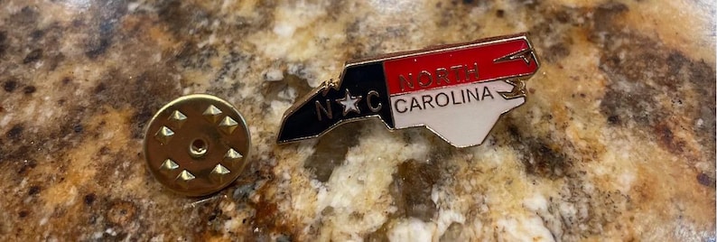 Puede incluir: Un pasador dorado y un pasador con la forma del estado de Carolina del Norte. El pasador presenta el contorno del estado en blanco, rojo y negro, con el texto "North Carolina" y "NC". El pasador es probablemente un recuerdo o un objeto de colecci&oacute;n.
