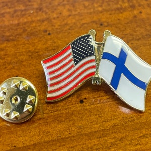 May include: A gold-colored pin with the American and Finnish flags side-by-side. The flags are enamelled with red, white, and blue for the American flag and blue and white for the Finnish flag.